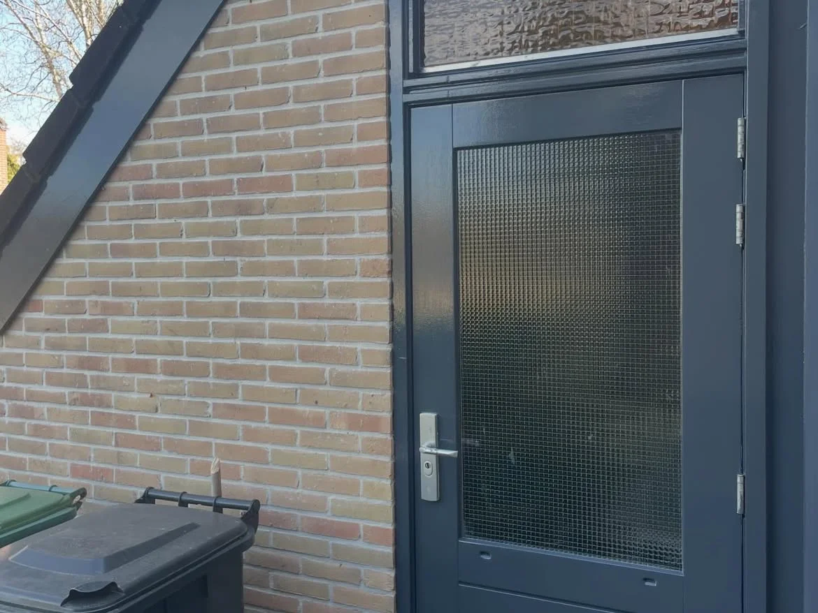 Een close-up van een moderne donkergrijze (antraciete) buitendeur tegen een lichtgele bakstenen muur. De deur heeft een groot rechthoekig raam gevuld met getextureerd ruitjespatroon privacyglas en is voorzien van een zilverkleurige deurkruk. Boven de deur bevindt zich een bovenlicht met vergelijkbaar mat glas. Links zijn de bovenkanten van groene en zwarte afvalcontainers zichtbaar bij de basis van de bakstenen muur.